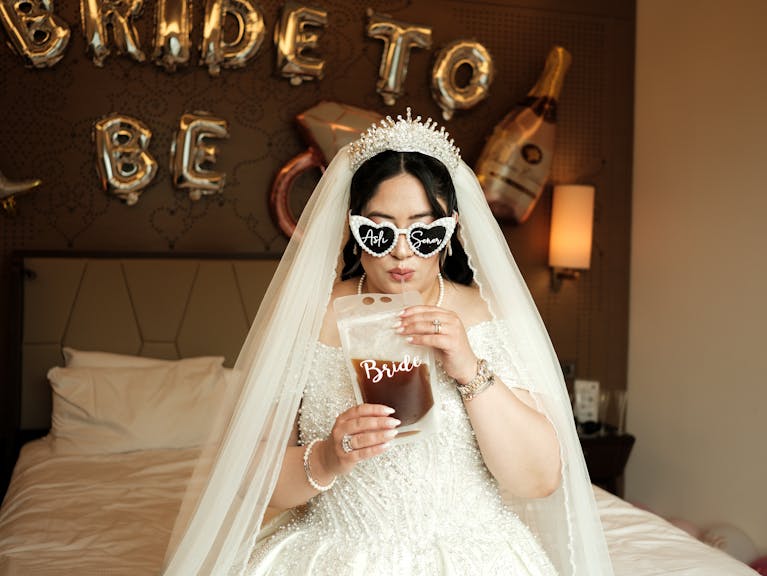 Bride in white dress and veil enjoys drink with fun party decor in the background.